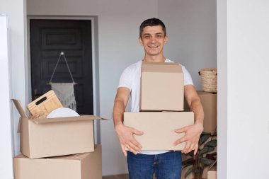 Beyaz tişört giyen mutlu bir adamın portresi. Elinde karton kutular, içinde eşyalar olan paketleri açıyor. Gülümseyerek kameraya bakıyor ve olumlu bir ifade sergiliyor..