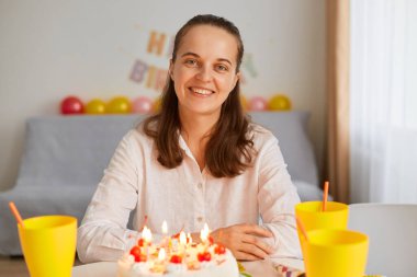 Beyaz elbiseli, beyaz elbiseli, neşeli pastayla masaya oturmuş, doğum gününü lezzetli bir tatlı ve mumla kutlayan, kameraya bakan, iyimser, iyimser bir kadının kapalı çekimleri..