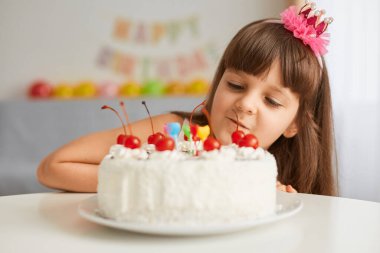 Tatlı, siyah saçlı, doğum günü pastasına bakan, misafirlerini bekleyen, arka planda balonlarla ve şenlikli yazıtlarla oturma odasında poz veren sevimli bir kız çocuk..