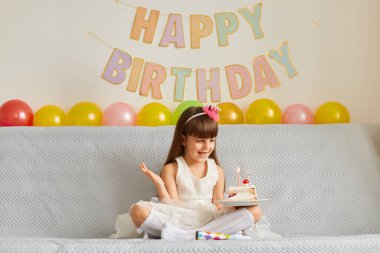 Doğum günü pastası ve yanan mumlarla kanepede oturan mutlu, gülümseyen küçük bir kızın yatay fotoğrafı. Bayramını kutluyor, kolunu kaldırıyor, mutluluğunu ifade ediyor..