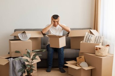 Beyaz tişört ve kot pantolon giyen, etrafı karton kutularla çevrili bir şekilde koltukta oturan, elleri kafasında poz veren, taşındıktan sonra hasar gören eşyalardan dolayı üzgün olan mutsuz bir adamın kapalı çekimleri..