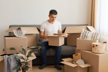 Beyaz tişört ve kot giyen üzgün adam karton kutularla çevrili koltukta oturuyor, dudak bükme ve keder ifadesiyle poz veriyor, paketin içine bakıyor, taşındıktan sonra kırılan şeylerden rahatsız oluyor..