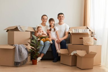 Taşındıktan sonra çocuklarla kanepede oturan, kameraya bakan, yeni bir daireye taşındıktan sonra mutluluklarını ifade eden, aileleri için ev alan gülümseyen çiftin portresi..