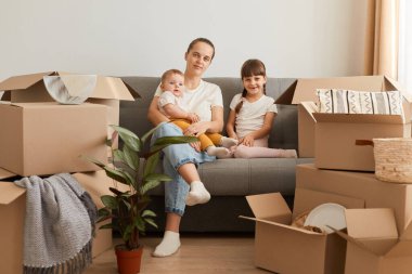 Kafkasyalı bir kadının, kızlarıyla kanepeye oturup kameraya bakarken, içinde eşyalar olan paketleri açarken, yeni bir daireye taşınırken,.