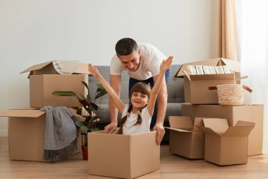 Playful father having fun in his new apartment on relocation day, happy man and his daughter playing together with carton box while moving, girl spreading hands, celebrating moving. clipart