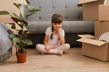 Çekici küçük bir kızın portresi. Etrafı karton kutularla çevrili, içinde eşyalar olan yerde oturuyor. Kameraya bakıyor, elini çenesinin altında tutuyor, yeni bir evde eşyalarını açıyor..