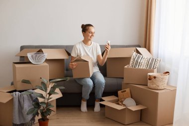 Beyaz tişört ve kot pantolon giyen, koltukta oturan, taşınma sırasında kutularla çevrili, akıllı telefonu elinde tutan, internette sörf yapan çekici bir kadın..