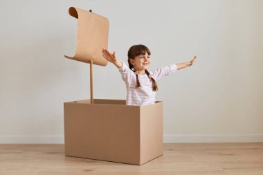 Kapalı mekanda, elinde kağıt bayrak, karton kutuda oturmuş, gemi kaptanı gibi poz veren, kollarını kaldırmış ve mutlu bir ifadeyle, denizdeymiş gibi davranan küçük bir kızın pozu..