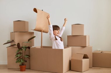 Heyecanlı küçük kız taşınma sırasında bayrakla karton kutudan gemi yapıyor ve yeni evde denizci rolü oynuyor, kollarını kaldırıyor, yeni daireye taşınmayı kutluyor..