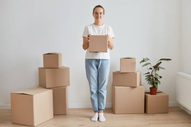 Etrafı karton kutularla çevrili, mutlu yüz ifadesiyle kameraya bakan mutlu genç bir kadının tam boy portresi..