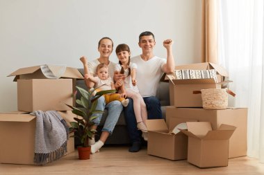 Yeni bir dairede öksüren mutlu bir aile, taşınmayı kutluyor, yumruklarını sıkmış, yeni bir eve taşınıyor, karton paketlerle çevrili..