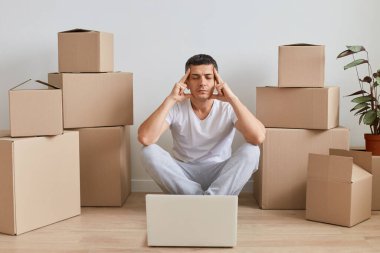 Kapalı alanda, tişört giyen yorgun bir adamın yerde kişisel eşyalarıyla karton kutuların yanında oturması, el ele tutuşması, baş ağrısı, taşınma sırasında online çalışmaktan bitkin düşmesi..