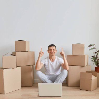 Beyaz tişörtlü esmer adamın, karton kutuların yanında otururken, bilgisayarın önünde poz verirken, yukarı doğru yükselirken, terfi için fotokopi çekerken,.