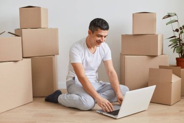 Beyaz tişört giyen mutlu bir gencin portresi karton kutuların yanında oturmuş, laptopta pozitif ve mutlu bir yüz ifadesiyle çalışıyor..