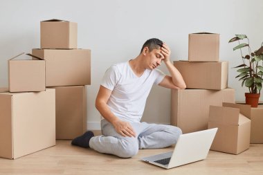 Beyaz tişörtlü yakışıklı, siyah saçlı bir adamın karton kutuların yanında otururken portatif bilgisayar üzerinde çalışırken çekilmiş yatay görüntüsü yorgun ve bitkin görünüyor..