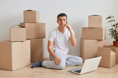 Üzgün, üzgün, beyaz tişörtlü bir adam resmi. Karton kutuların yanında, kişisel yığınla oturmuş, telefonla konuşurken kameraya bakıyor, defter üzerinde çalışıyor..