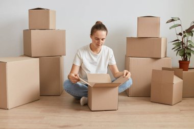 Beyaz tişörtlü, beyaz tişörtlü, beyaz saçlı beyaz bir kadının karton kutuların yanında oturduğu, taşınma sırasında kişisel eşyalarının olduğu paketleri açtığı yatay bir fotoğraf..