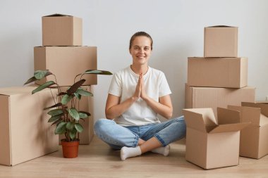 Beyaz tişörtlü güzel bir kadın karton kutuların yanında oturmuş nilüfer pozu veriyor, avuç içlerini bir arada tutuyor, dua ediyor ya da yoga yapıyor..