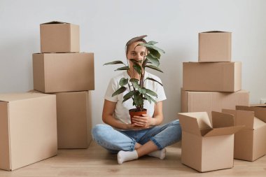 Yatay olarak beyaz tişört giyen bir kadın karton kutuların yanında oturmuş elinde çiçekle çiçek saksısı, yüzünü çiçeklerle örtüyor..