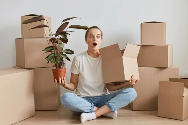 Indoor shot of astonished woman wearing white T-shirt sitting on the floor near cardboard boxes with stuff, carrying flower and carton box, keeps mouth opened, expressing astonishment.