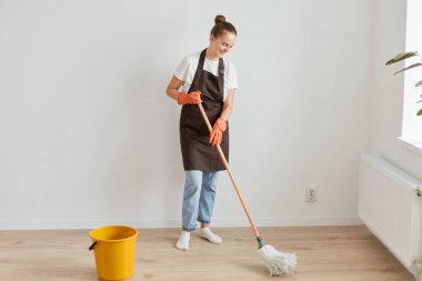Turuncu lastik eldiven, kahverengi önlük ve kot pantolon giyen, paspas tutan, iyimser duygularını ifade eden pozitif, neşeli ev kadınlarının tam boy fotoğrafı..