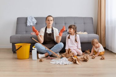 Evin içinde kahverengi önlük, kot pantolon ve beyaz tişört giyen bir kadın evini çocuklarla temizlerken, yorgun ve bitkin düşmüşken yoga pratiğiyle rahatlamaya çalışırken..