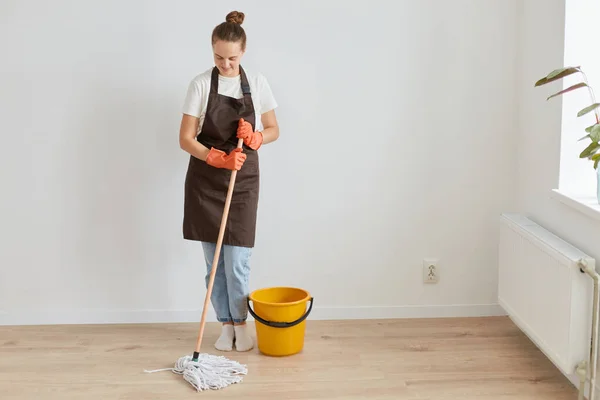 Full length portrait of woman wearing orange rubber gloves, brown apron and jeans washing floor at home, looking smiling down, doing household chores during weekend.
