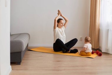Genç annenin kapalı alanda bebeğiyle birlikte fiziksel yoga egzersizleri yaparken kollarını kaldırmış, avuç içlerini bir arada tutarak, rutin çalışma günlerinde dinlenirken..