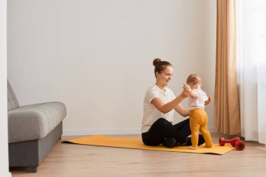 Atletik kadın nilüfer çiçeğinin altında, pencerenin önündeki koltuğun yanında poz veriyor, beyaz tişört ve siyah tozlar giyiyor, küçük kızlarının ellerini tutuyor, bebekle spor yapıyor..