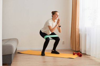 Beyaz tişörtlü, siyah tozlu çekici bir kadının uzun boylu portresi, elastik bantla mekik çekmek, evde egzersiz yapmak, fitness ve sağlıklı yaşam tarzı..