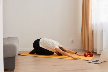 Kapalı alanda spor yapan bir kadının yoga yaparken, vücudunu esnerken, beyaz tişört ve siyah tozluk giyerken, aktif sağlıklı yaşam tarzında, evde egzersiz yaparken..