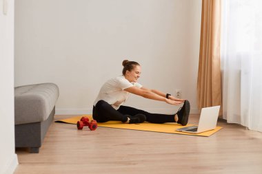 Bacakları açık, elleri esnek, spor esneme egzersizleri yapan, beyaz tişört ve siyah tayt giyen, evde spor yapan, paspasın üzerinde oturan sağlıklı jimnastikçi kadın..