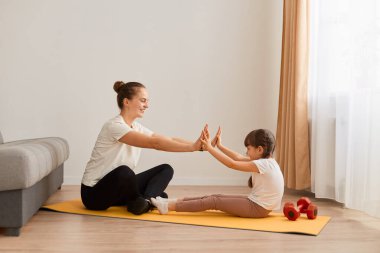 Saçları topuzlu çekici bir kadının, küçük kızıyla spor yaptığı, yoga minderinde oturduğu ve avuçlarına dokunduğu, evde birlikte spor yaptığı görüntüsü..