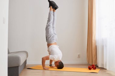 Kapalı alanda çekilen, siyah saçlı, kanepede baş aşağı duran, yoga, akrobatik aktiviteler ve evde beyaz tişört ve gri pantolon giyen spor adamı..