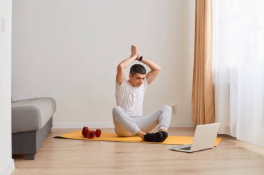 Atletik bir adamın yoga yapması, gündüz evde eğitim videosu izlemesi, dua ederek kollarını kaldırması, dizüstü bilgisayara bakması, evde antrenman yapması..