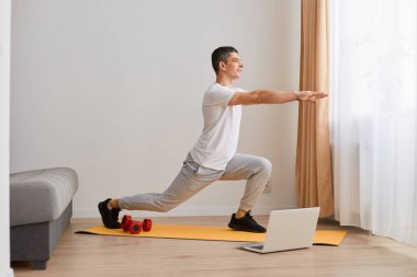 Kişisel antrenör. Erkek fitness eğitmeninin evde dizüstü bilgisayar kullanarak antrenman yaparken vücudunu esnetirken tam boy görüntüsü..