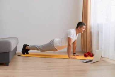 Spor giyim kuşamında siyah saçlı sıska bir adamın kapalı çekimleri. Yoga yaparken, düz kollar üzerinde Phalakasana egzersizi yaparken, kasları çalıştırırken. Daha yüksek tahta duruşu.