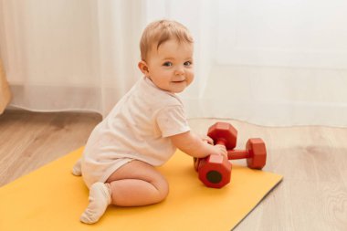 Kapalı alanda, yoga minderinde oturan sevimli bir çocuğun halterli kameraya gülümseyerek baktığı, spor malzemelerini keşfettiği bir fotoğraf..