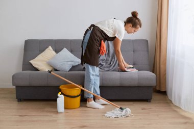 Genç bir kadının evini temizlerken, kot pantolon ve kahverengi önlük giyerken, ev hanımı pencerenin yanındaki aydınlık oturma odasında ev işlerini yaparken yan görüntüsü..