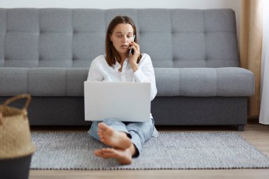 İç mekanda, kot pantolon ve beyaz gömlek giyen, dizüstü bilgisayarında çalışan ya da akıllı telefondan çalışıp konuşan, şaşkınlığını dile getiren, şaşırmış çekici bir kadının fotoğrafı..