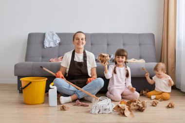 Beyaz tişört, kahverengi önlük ve kot pantolon giyen, evi temizleyen, çocuklarıyla yerde oturan, kameraya bakan, ev işlerini yaparken dinlenen bir kadının yatay görüntüsü..