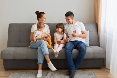 Mutlu bir aile oturma odasında kanepede rahat kıyafetler giyer, hafta sonunu birlikte geçirir, cep telefonlarını kullanır, olumlu duygular ifade eder, birlikte zaman geçirmekten memnun olur..