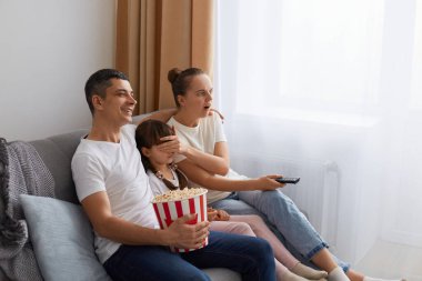 Çift kanepede oturup birlikte film izlerken, anne kızının gözlerini kapatır ve ağzı açık bakar, baba gülümser, aile birlikte vakit geçirir..