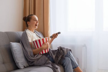 Çekici bir kadının portresi. Bun saç stili olan çekici bir kadın. Battaniyeye sarılmış bir şekilde kanepeye oturuyor ve lezzetli atıştırmalıklarla film seyrediyor..