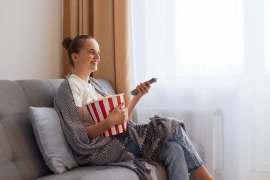 Bun saçlı bir kadının gri battaniyeye sarılı olarak kanepeye oturup film izlemesi ve patlamış mısır yemesi, ilginç bir film ya da televizyon programının keyfini çıkarması..