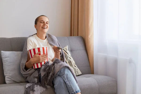 Indoor shot of woman sitting on sofa wrapped in plaid and watching funny movie and eating popcorn, spending her weekend alone, enjoying comedy film or tv show.