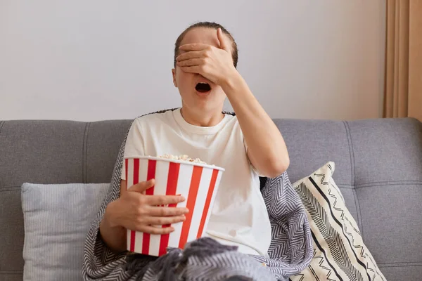 Beyaz tişört giyen, kanepeye oturup patlamış mısırla kağıt kovayı kucaklayan, tek başına korku filmi seyreden, avuçlarıyla gözlerini kapayan korkmuş bir kadının kapalı çekimi..