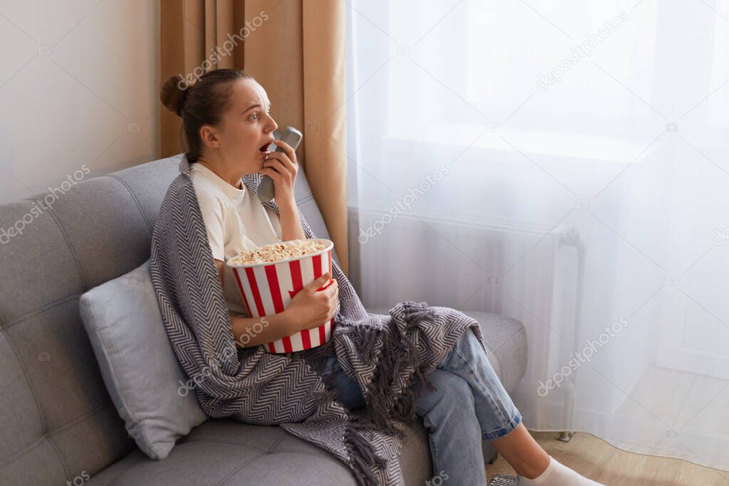 Profile portrait of of attractive socked scared woman with bun hairstyle sitting on couch wrapped in blanket and watching scary movie, embracing bucket with popcorn, keeps mouth open.