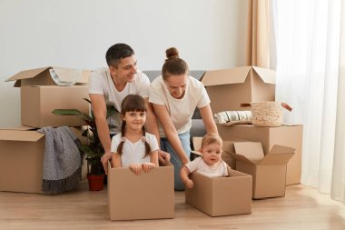 Yeni bir eve taşınırken beyaz t-shirtler giyip, aileleri karton kutularda çocuklarıyla oynarken birlikte yerde oturup eğlenen mutlu beyaz bir çift..