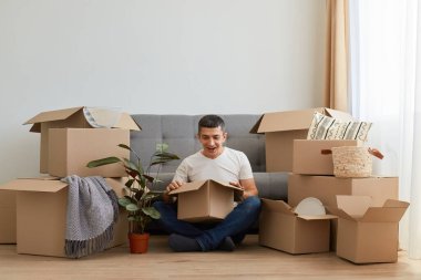 Beyaz t-shirt ve kot giyen, elinde karton kutuyla yerde oturan, şaşırmış yüz ifadesiyle içeri bakan, kişisel eşyalarını boşaltan şaşırmış bir adamın görüntüsü..